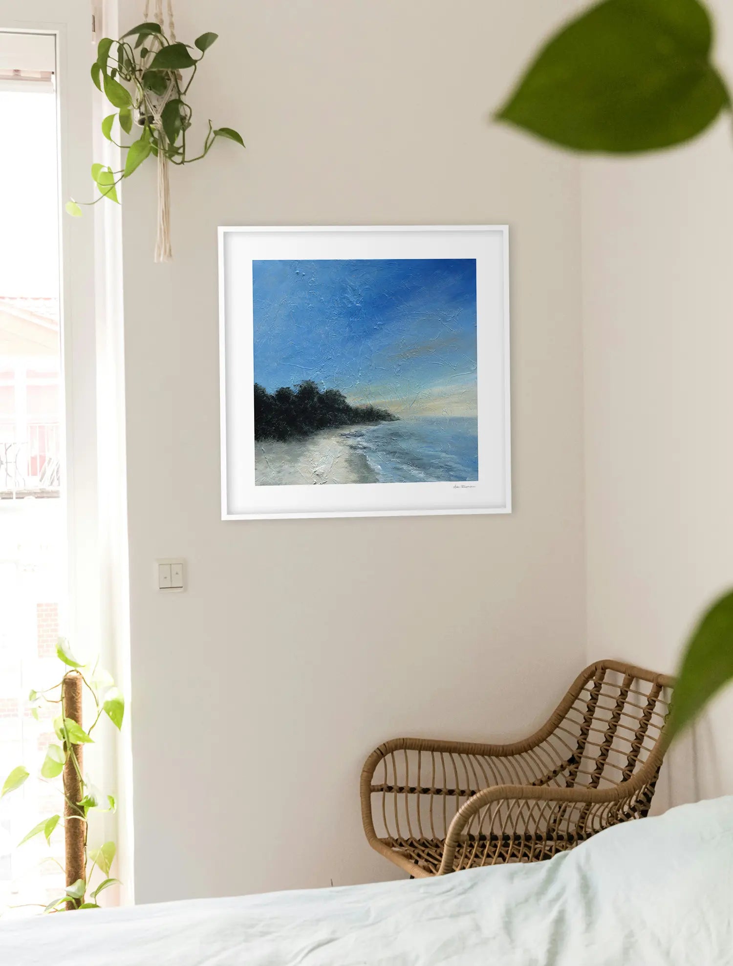 Framed artwork of a beach scene on a wall with a wicker chair and plants in the room.