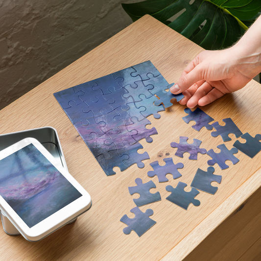 A hand is placing a jigsaw puzzle piece onto a completed square puzzle showing a purple and blue landscape. The puzzle pieces are scattered on a light wood table next to a white smartphone displaying the puzzle design and a small metal tin.