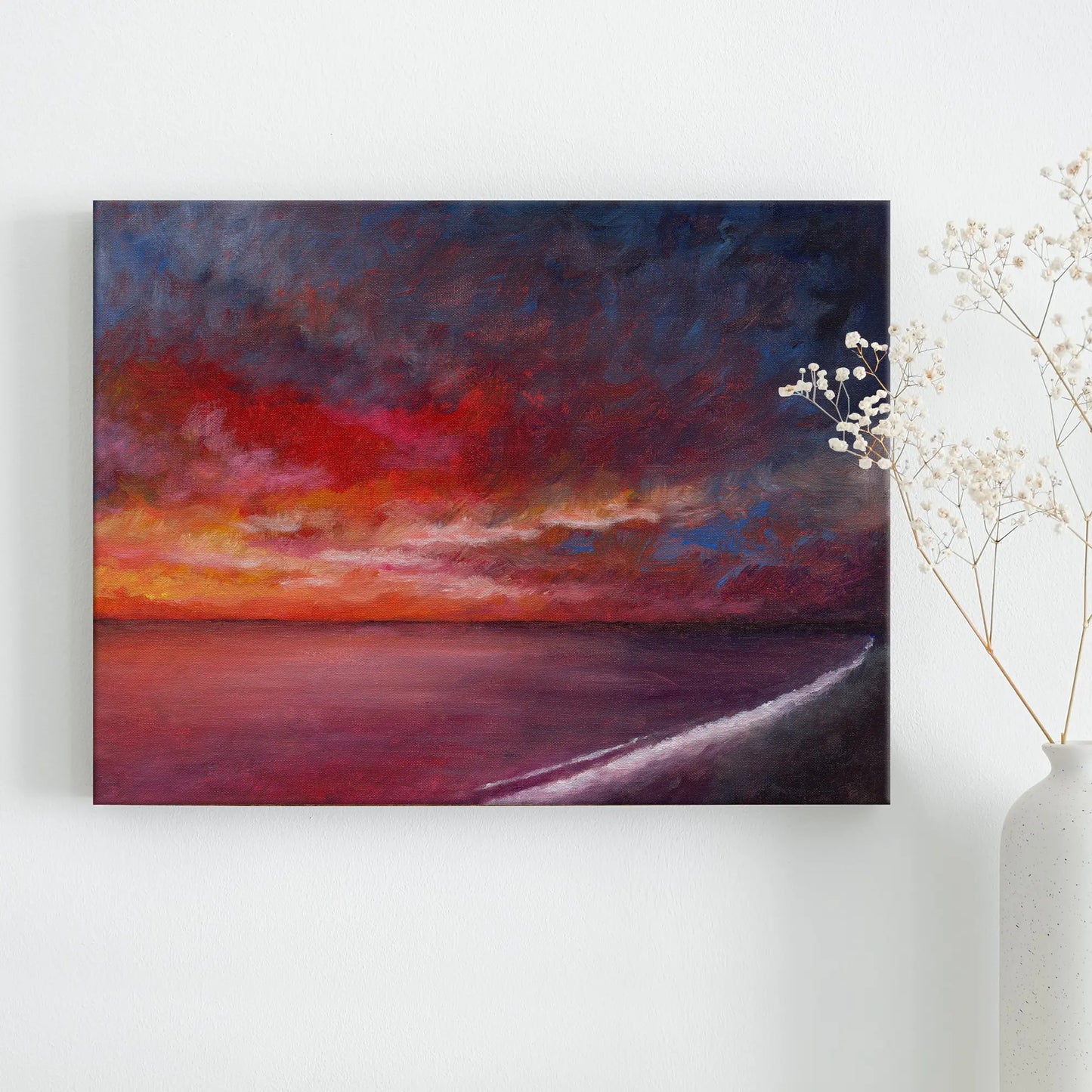 Painting of a sunset over water on a white wall with a vase of dried flowers.