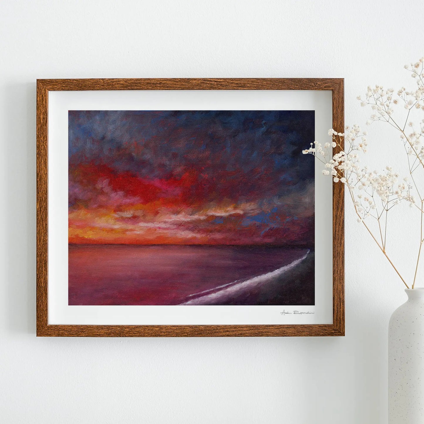 Framed painting of a sunset over water on a white wall with a vase of flowers.