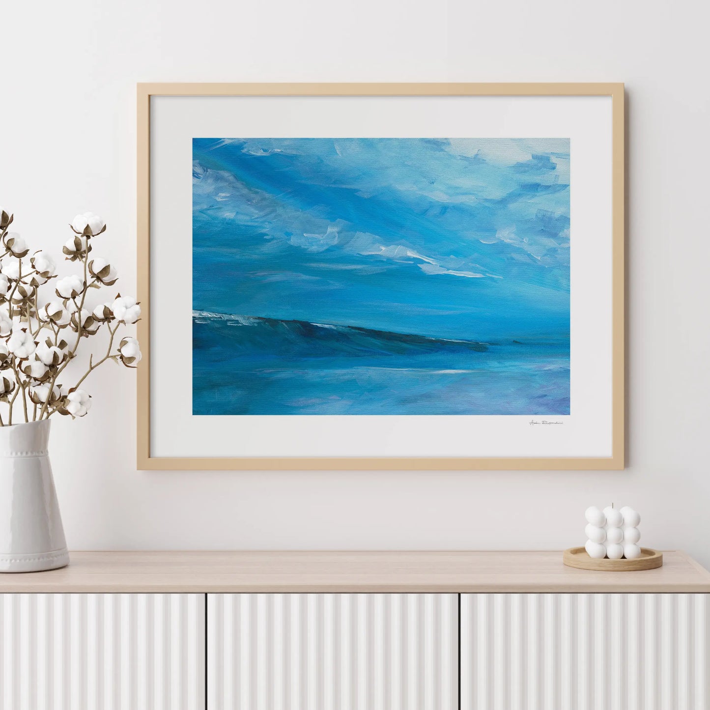 Framed artwork of a blue sky and ocean scene on a white wall with decorative items.